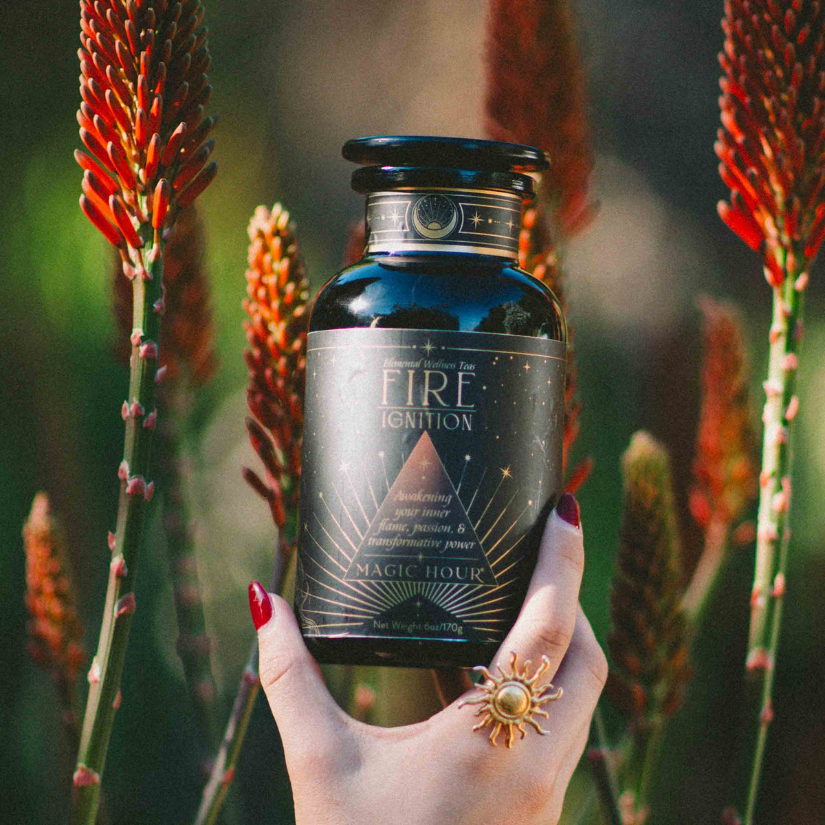 A hand with red nails and a sun-shaped ring holds a dark bottle labeled “Fire Ignition” amid tall orange flowers and green stems in soft sunlight, evoking the energizing blend of Fire: Spiced Black Tea with Ginger and Cardamom to Ignite Courage and Vitality.
