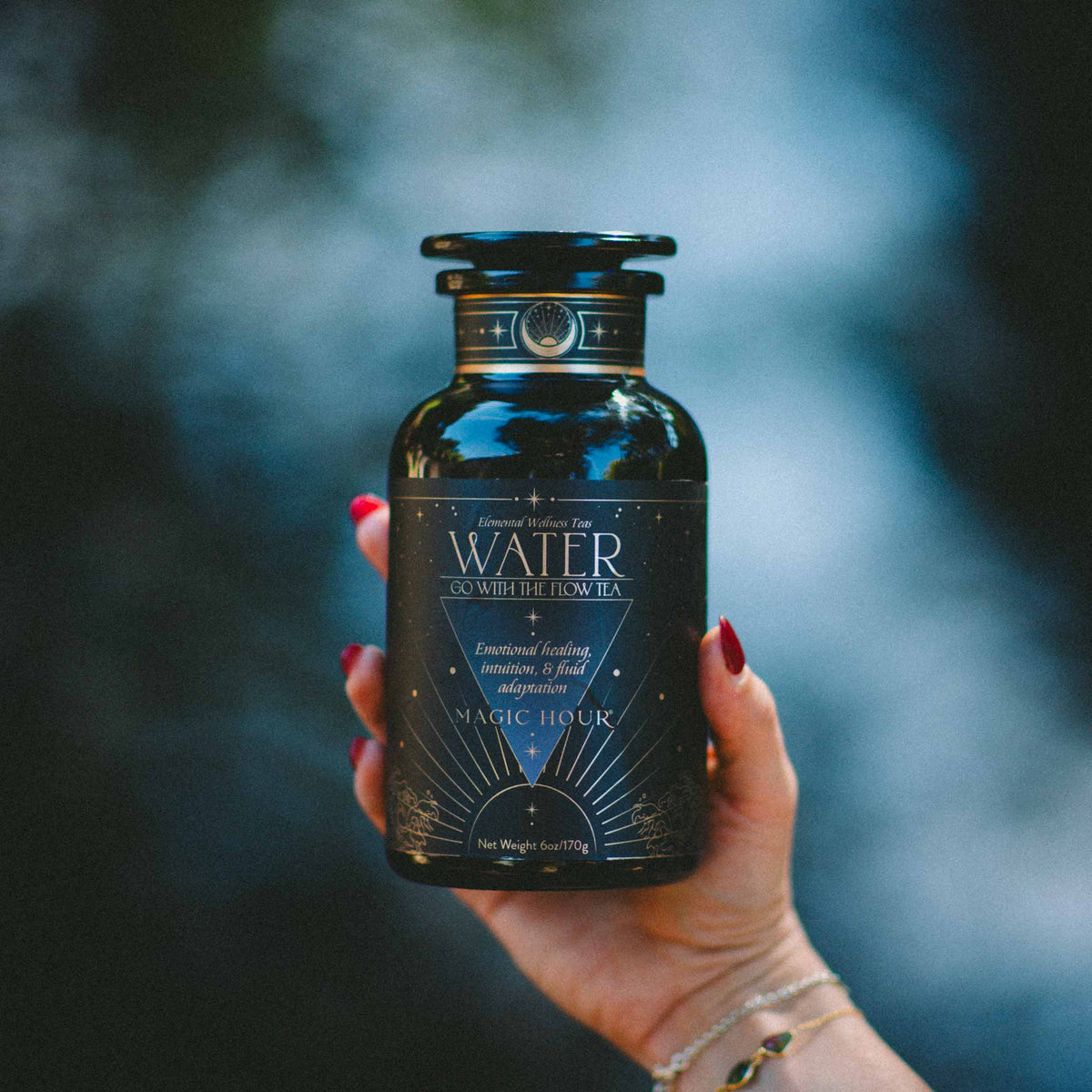 A hand with red nail polish holds a dark glass jar labeled “Water White Tea: Soothing Lemon Balm Rose Reishi Tea for Going with the Flow” by Magic Hour, featuring a calming herbal tea blend with reishi mushroom, against a blurred outdoor background.