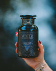 A hand with red nail polish holds a dark glass jar labeled “Water White Tea: Soothing Lemon Balm Rose Reishi Tea for Going with the Flow” by Magic Hour, featuring a calming herbal tea blend with reishi mushroom, against a blurred outdoor background.