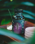 A dark glass bottle labeled "Aphrodite's Alchemy: Damiana & Rose White Tea" with a purple floral design sits beside a pink cup on wood, surrounded by green leaves and soft light, evoking the enchanting allure of Greek mythology.
