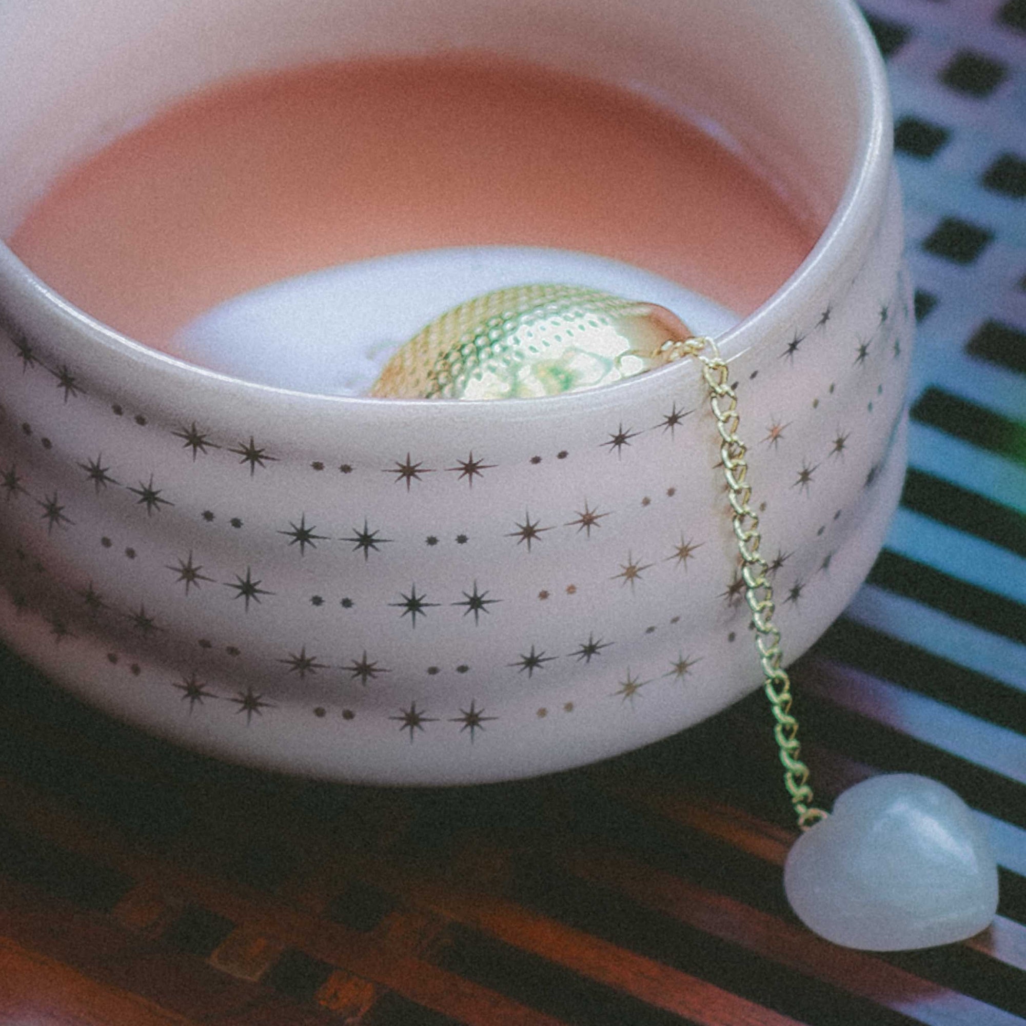 A creamy pink drink fills a blush cup adorned with gold stars, while the Rose Quartz Heart Tea Strainer—with its delicate chain and heart-shaped charm—rests gracefully inside. This elegant tool is perfect for elevating your tea ritual, as the cup sits atop a patterned surface, inviting tranquil moments inspired by rose quartz serenity.