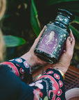 A person with wavy hair holds a jar of "Aphrodite's Alchemy: Damiana & Rose White Tea," featuring an Aphrodite-inspired illustrated label, surrounded by green leafy plants.