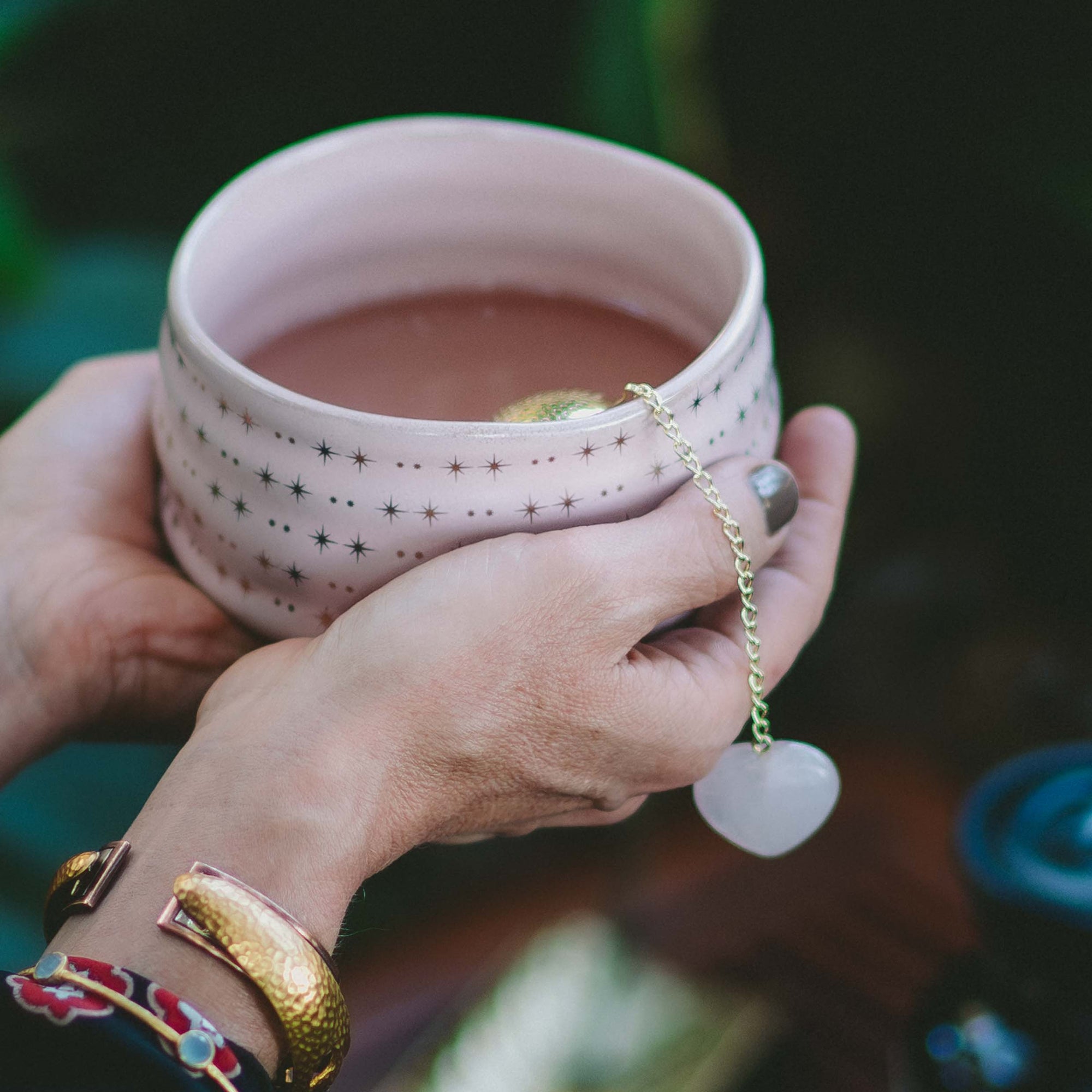 A person cradles a pink, star-patterned mug of tea, savoring their daily ritual with a heart-shaped infuser. Wearing a gold bracelet and beautifully painted nails, they celebrate self-love with every sip—especially as the Rose Quartz Heart Tea Strainer brings a delicate, uplifting charm to their moment of calm.
