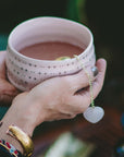 A person cradles a pink, star-patterned mug of tea, savoring their daily ritual with a heart-shaped infuser. Wearing a gold bracelet and beautifully painted nails, they celebrate self-love with every sip—especially as the Rose Quartz Heart Tea Strainer brings a delicate, uplifting charm to their moment of calm.