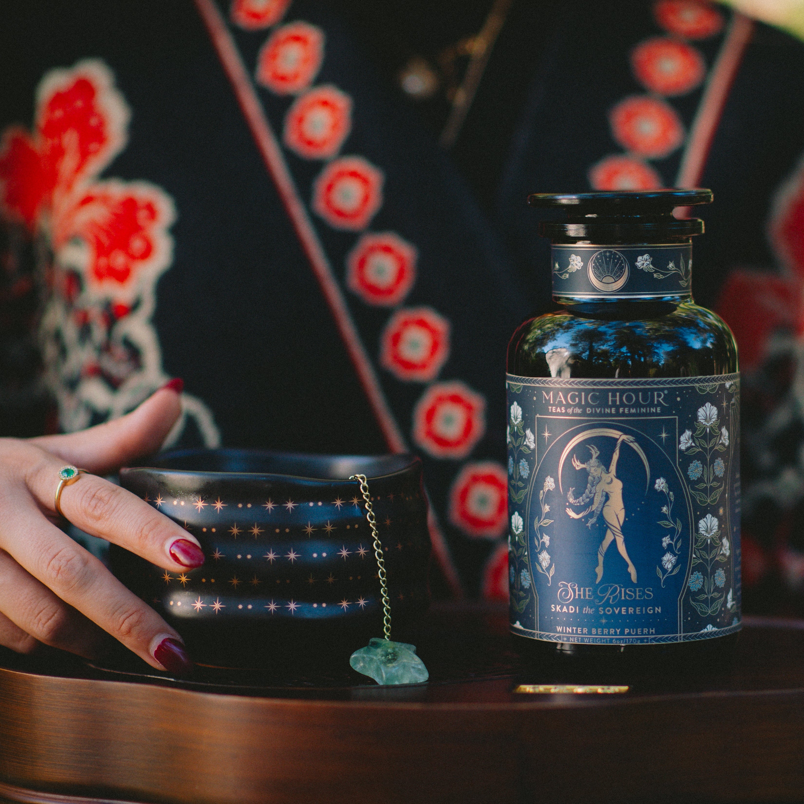 A person with painted nails and a turquoise ring holds a black mug with star patterns. Beside it sits a 