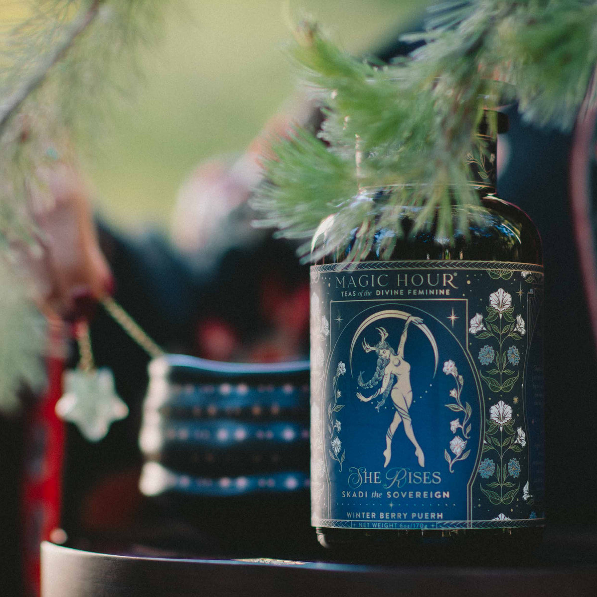 A bottle of “Magic Hour” tea labeled “Skadi The Sovereign: Winter Berry Puerh” sits on a table beside a hand holding a star-shaped object and a stack of dark cups. Pine branches frame the scene, evoking Skadi’s sovereignty and the spirit of winter resilience.