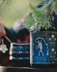 A bottle of “Magic Hour” tea labeled “Skadi The Sovereign: Winter Berry Puerh” sits on a table beside a hand holding a star-shaped object and a stack of dark cups. Pine branches frame the scene, evoking Skadi’s sovereignty and the spirit of winter resilience.