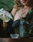 A person pours decaf green tea from a glass teapot into a small cup beside a dark jar labeled “Air: Mint, Mullein & Citrus Tea for Respiratory Support” on a wooden surface, with greenery in the background, evoking a sense of calm and respiratory wellness.