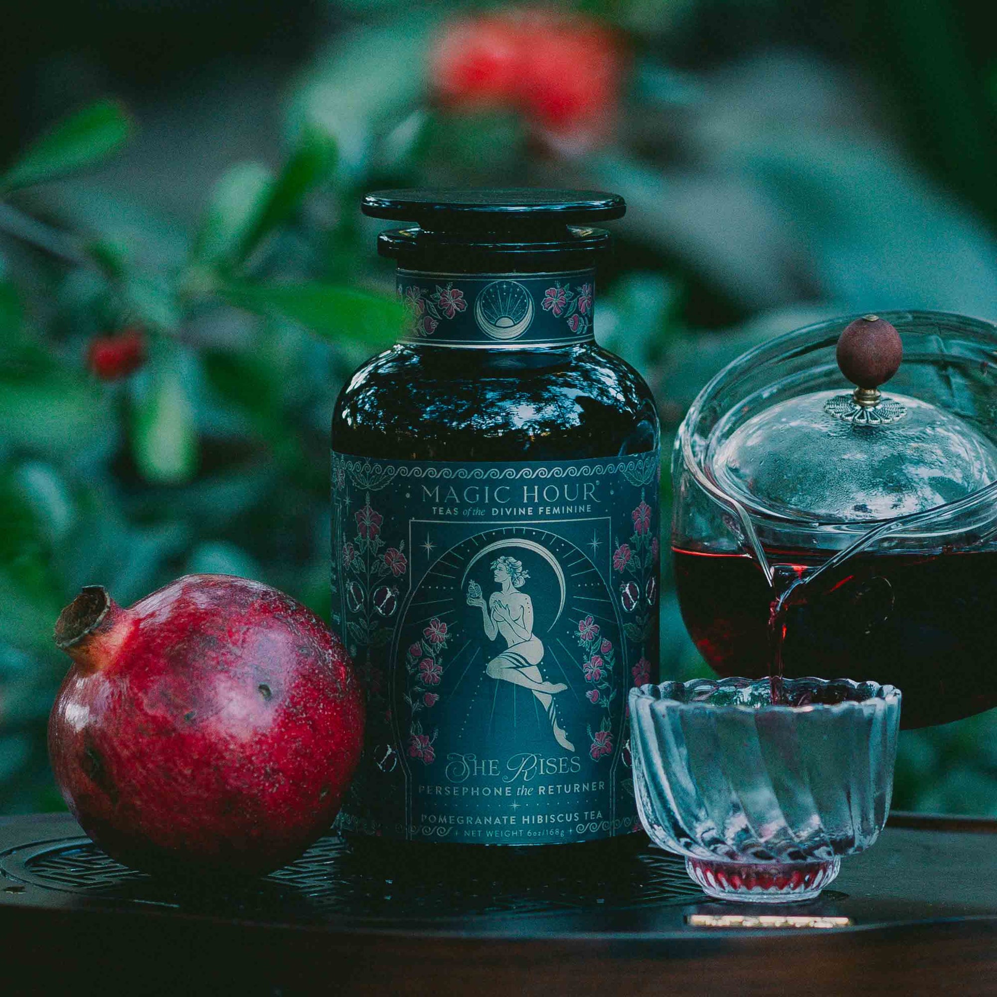 A black jar labeled "Monthly Magic Tea Subscription Box" stands by a whole pomegranate, as a glass teapot pours red tea into a cup—inviting you to a ceremonial tea experience outdoors among lush green foliage.