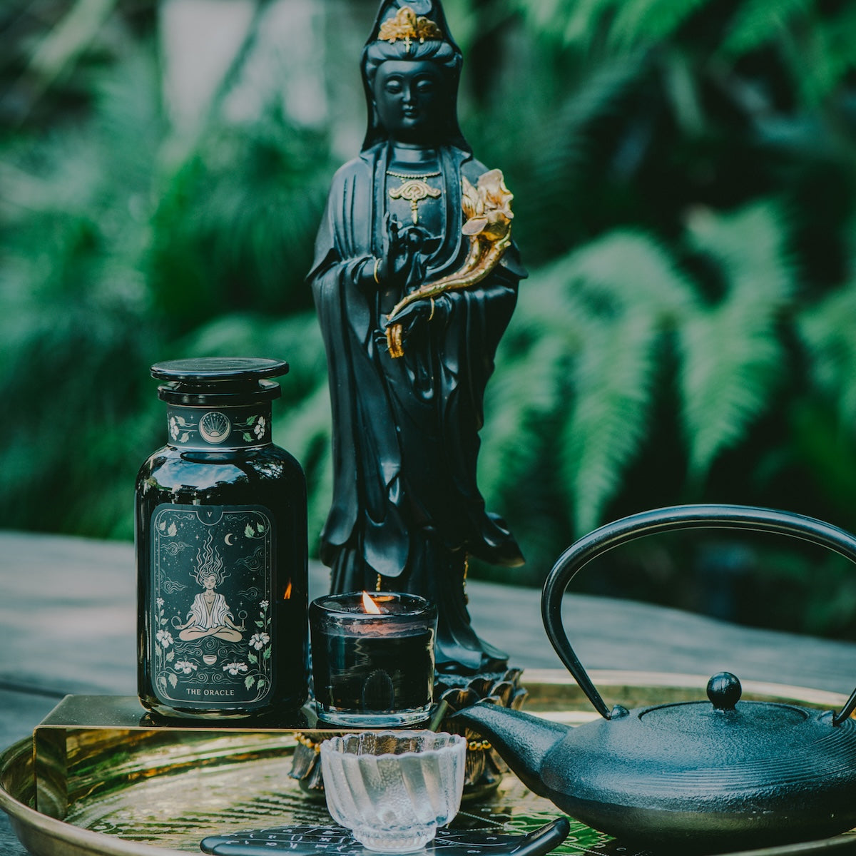 A black statue stands on a tray with an ornate black jar, a small candle in a holder, and a black teapot. The background features lush greenery, creating a serene atmosphere perfect for enjoying Magic Hour&#39;s Monthly Magic First Sips Tea Subscription Box.