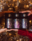 Two pairs of hands hold a She Rises Goddess Moonlit Bloom Apothecary Gift Box containing three dark glass apothecary jars of moonlight white tea, each featuring ornate, illustrated labels.