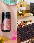 A collage showcasing a Diamond April Birthstone Mini Gift Set, which includes a tea tin decorated with a gemstone design. Alongside it is an Aquamarine Wellness Gift Set. A Magic Hour tea box with a celestial pattern stands next to an Opalite Moon Tea Strainer on a reflective surface. The background is enhanced with flowers, pink cloth, and decorative elements.