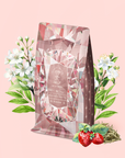 A pink, diamond-patterned package of Diamond - Champagne & Strawberry Jasmine White Tea for Beautiful Skin stands upright, adorned with white floral designs and surrounded by jasmine flowers, green leaves, organic strawberries, and loose Silver Needle White Tea leaves—all arranged on a pale pink background.