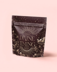 A resealable tea package labeled “Divine Timing Vitality” displays celestial and botanical art—stars, flowers, a bird—symbolizing the adaptogenic tea's natural benefits, set on a deep brown backdrop atop a light pink surface.