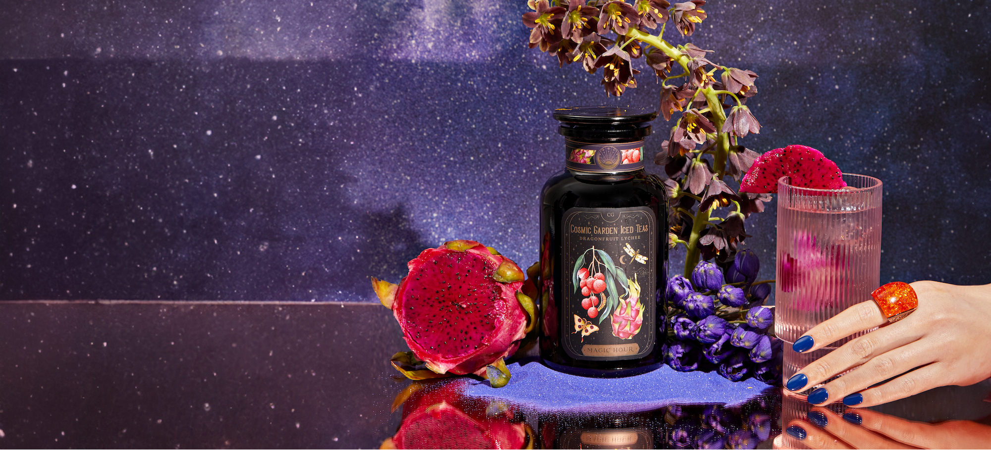 A jar of Twinings Cosmic Cranberry & Lime tea is surrounded by dragon fruits, a glass of tea, and purple flowers against a starry galaxy backdrop. A hand with navy blue nail polish and a red ring holds the glass of tea.