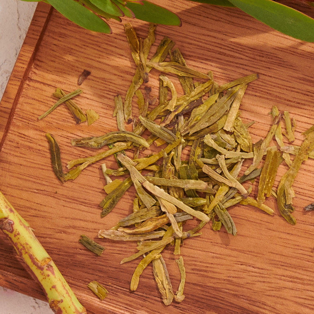 Organic Dragonwell Longjing Green Tea from ARCHIVE is beautifully displayed on a wooden surface, accompanied by small green leaves and a piece of tree bark. With every brew, this premium Longjing tea captures the essence of Lin'an County right in your cup.