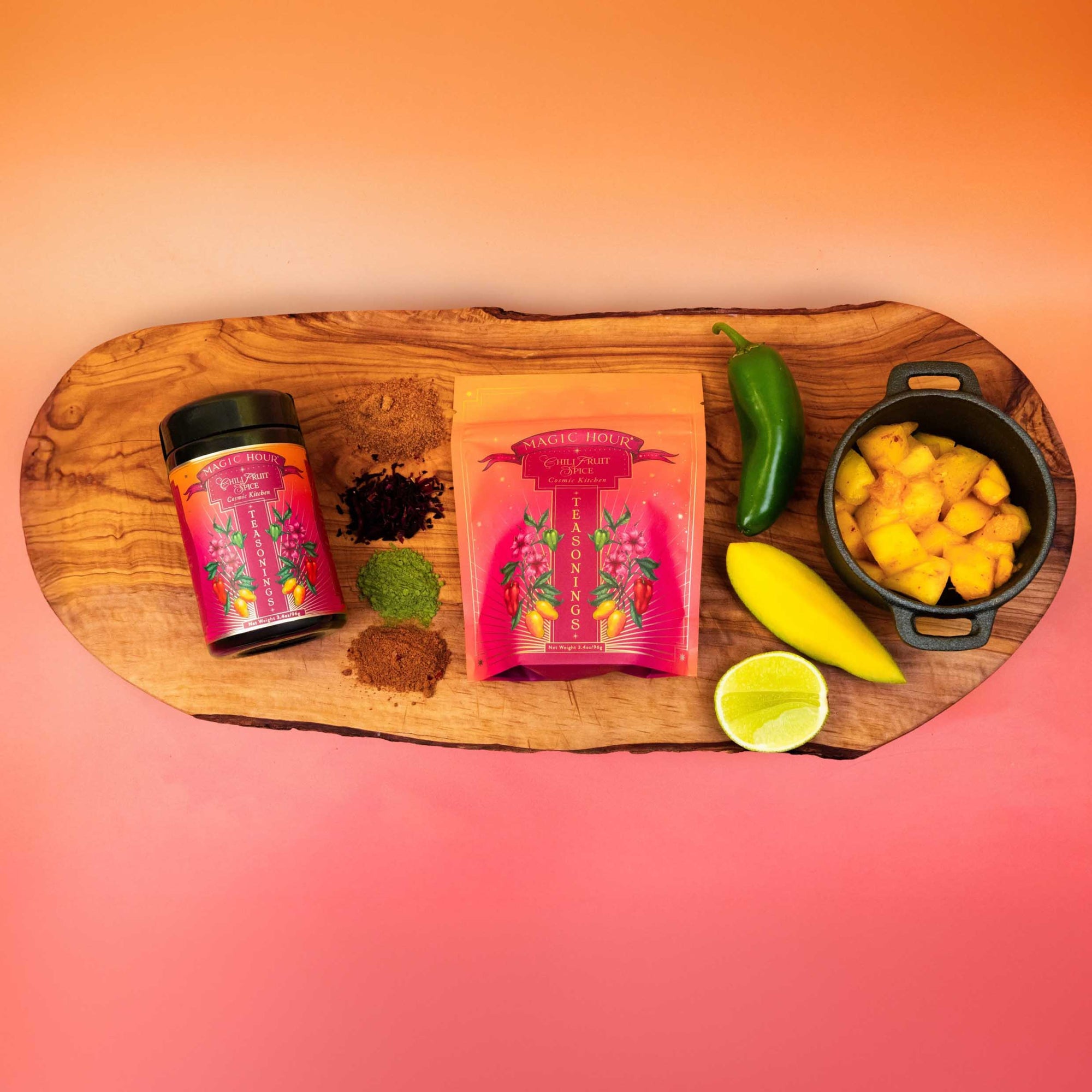 A wooden board displays a jar and pouch of Teasoning: Chili Fruit Spice tea, surrounded by loose tea, matcha hibiscus, jalapeño, yellow chile, pineapple chunks, halved lime, and tropical seasoning on an orange-to-pink gradient backdrop.