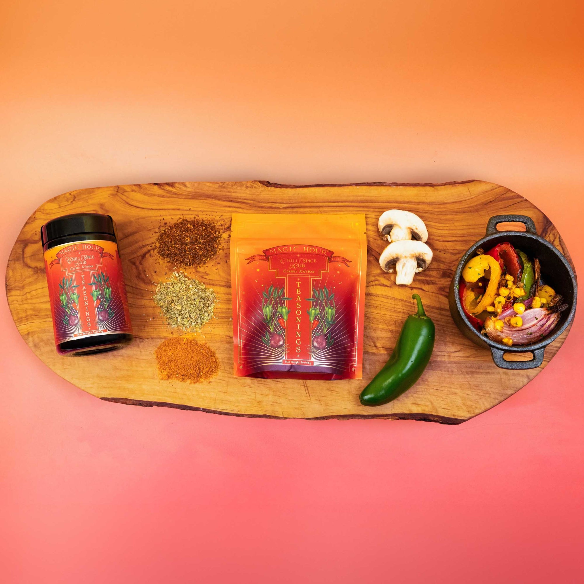 A wooden board displays a jar and pouch of Teasoning: Chili Spice Rub, four spice piles including an umami blend, a jalapeño, mushrooms, and a bowl of vibrant cooked veggies, all set against an orange gradient background.