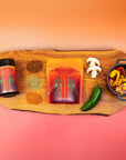 A wooden board displays a jar and pouch of Teasoning: Chili Spice Rub, four spice piles including an umami blend, a jalapeño, mushrooms, and a bowl of vibrant cooked veggies, all set against an orange gradient background.