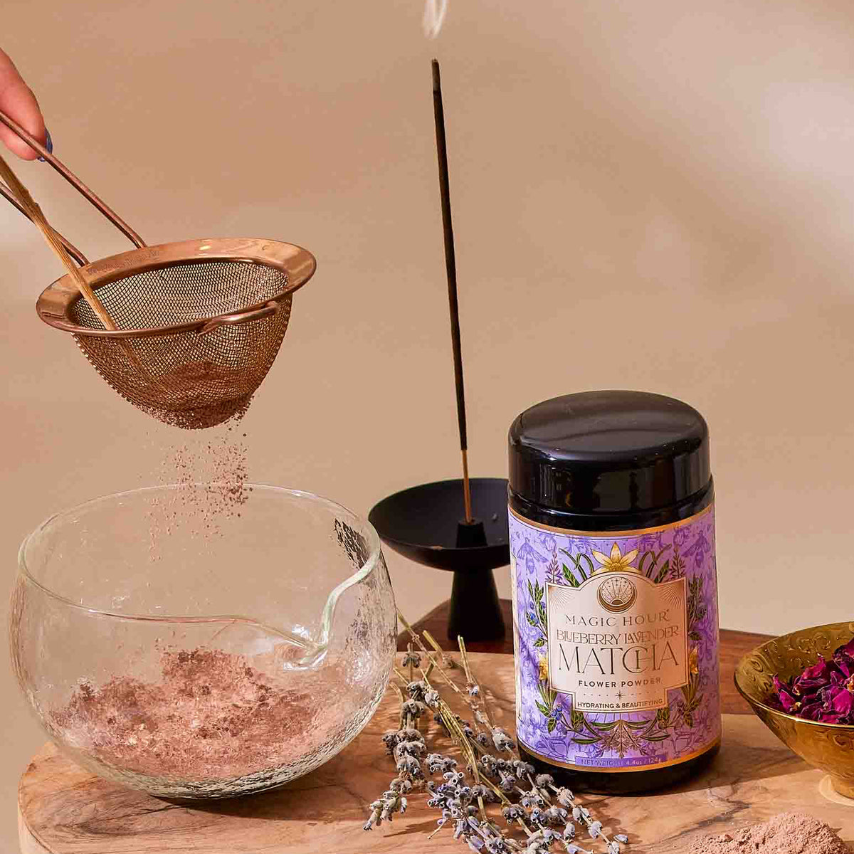 A jar of Flower Powder: Blueberry Lavender Matcha sits on a wooden board with a whisk, dried flowers, a teapot, cup of tea, sieve in use, and calming incense on a sunlit wooden table.