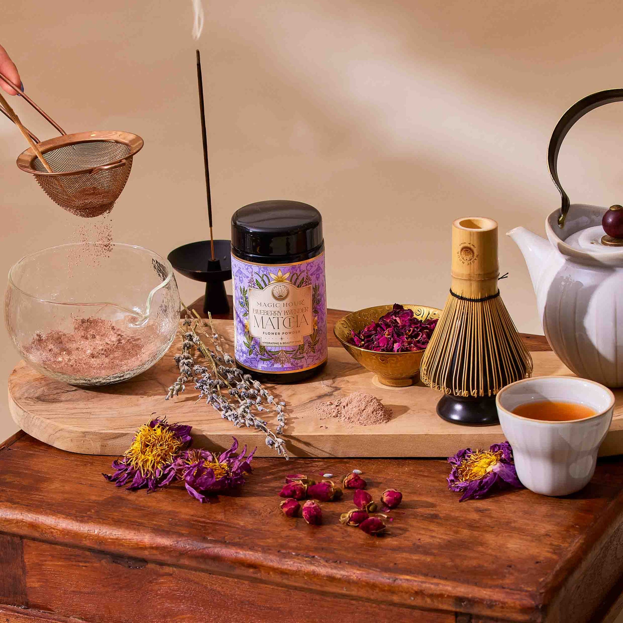 A jar of Flower Powder: Blueberry Lavender Matcha sits on a wooden board with a whisk, dried flowers, a teapot, cup of tea, sieve in use, and calming incense on a sunlit wooden table.