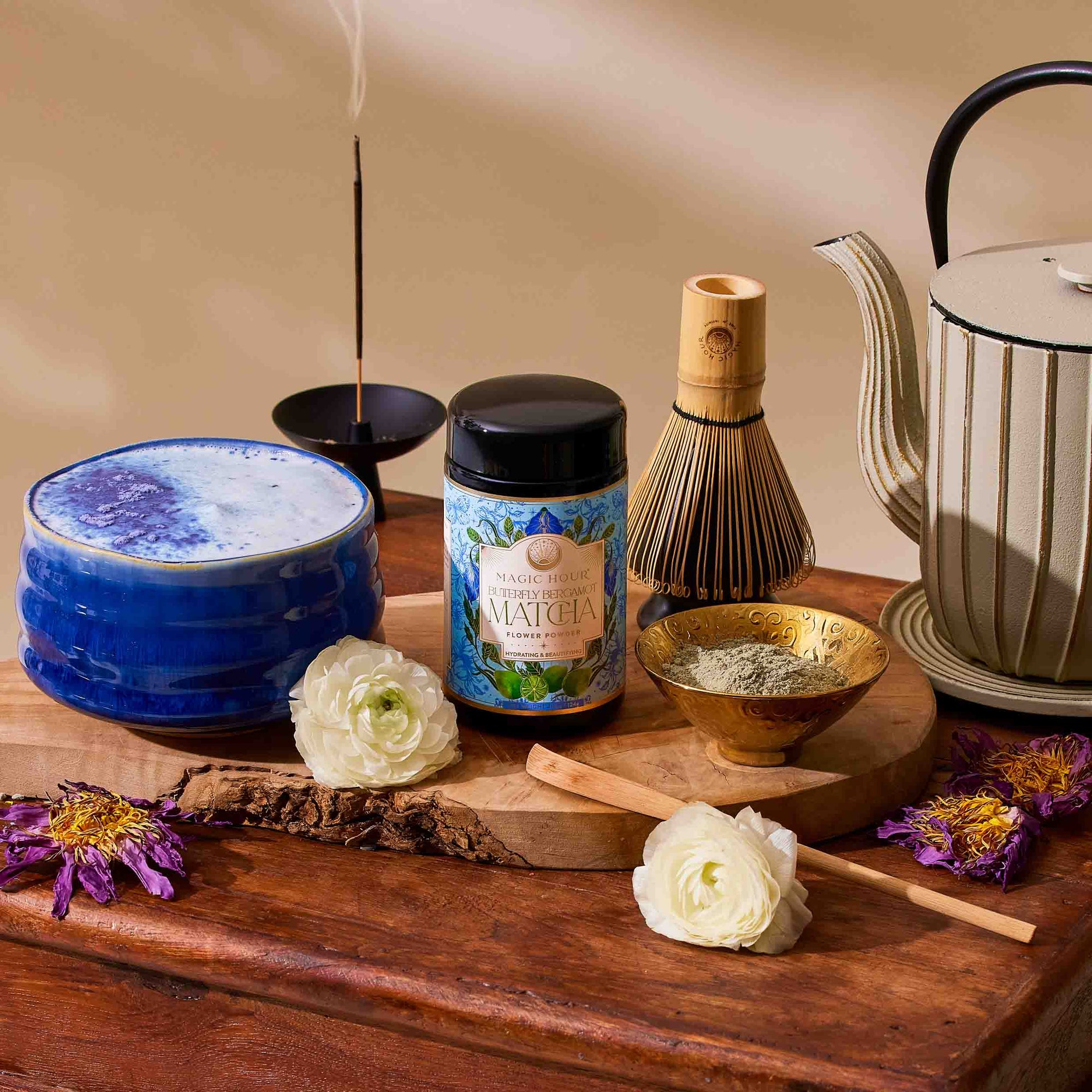 The Flower Powder: Butterfly Bergamot Matcha set features a blue bowl, ceremonial matcha canister, gold matcha powder bowl, bamboo whisk, teapot, incense stick, and white flowers on a wooden surface.