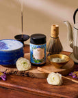 The Flower Powder: Butterfly Bergamot Matcha set features a blue bowl, ceremonial matcha canister, gold matcha powder bowl, bamboo whisk, teapot, incense stick, and white flowers on a wooden surface.