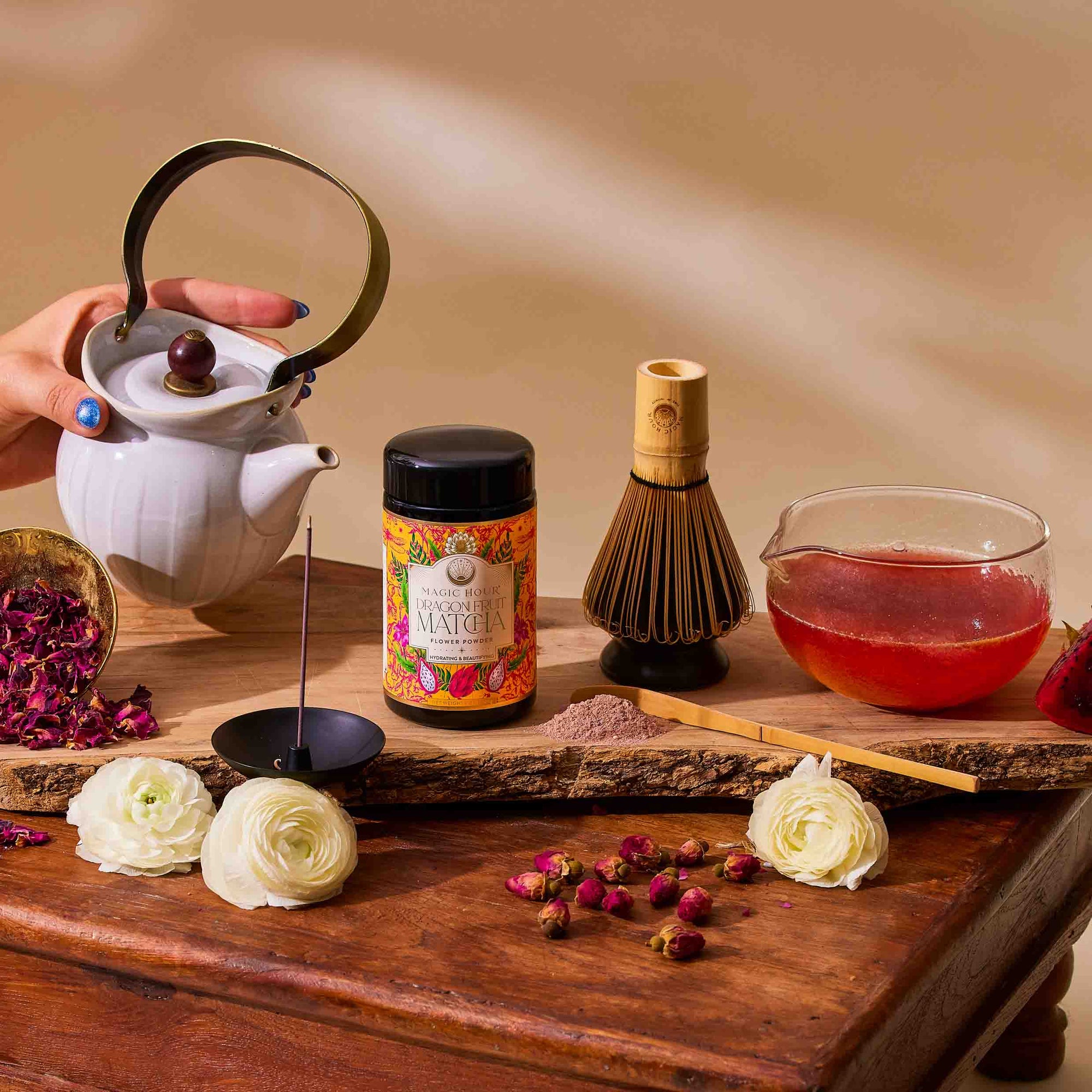 A hand pours tea from a white teapot beside the Flower Powder: Matcha Bundle, including ceremonial matcha, a bamboo whisk, tea bowl, rose buds, flowers, and tea tools on a rustic wooden surface.