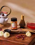 A hand pours tea from a white teapot beside Flower Powder: Dragon Fruit Matcha, a bamboo whisk, glass of red tea, white flower, matcha scoop rich in vitamin C, and rosebuds on wood.