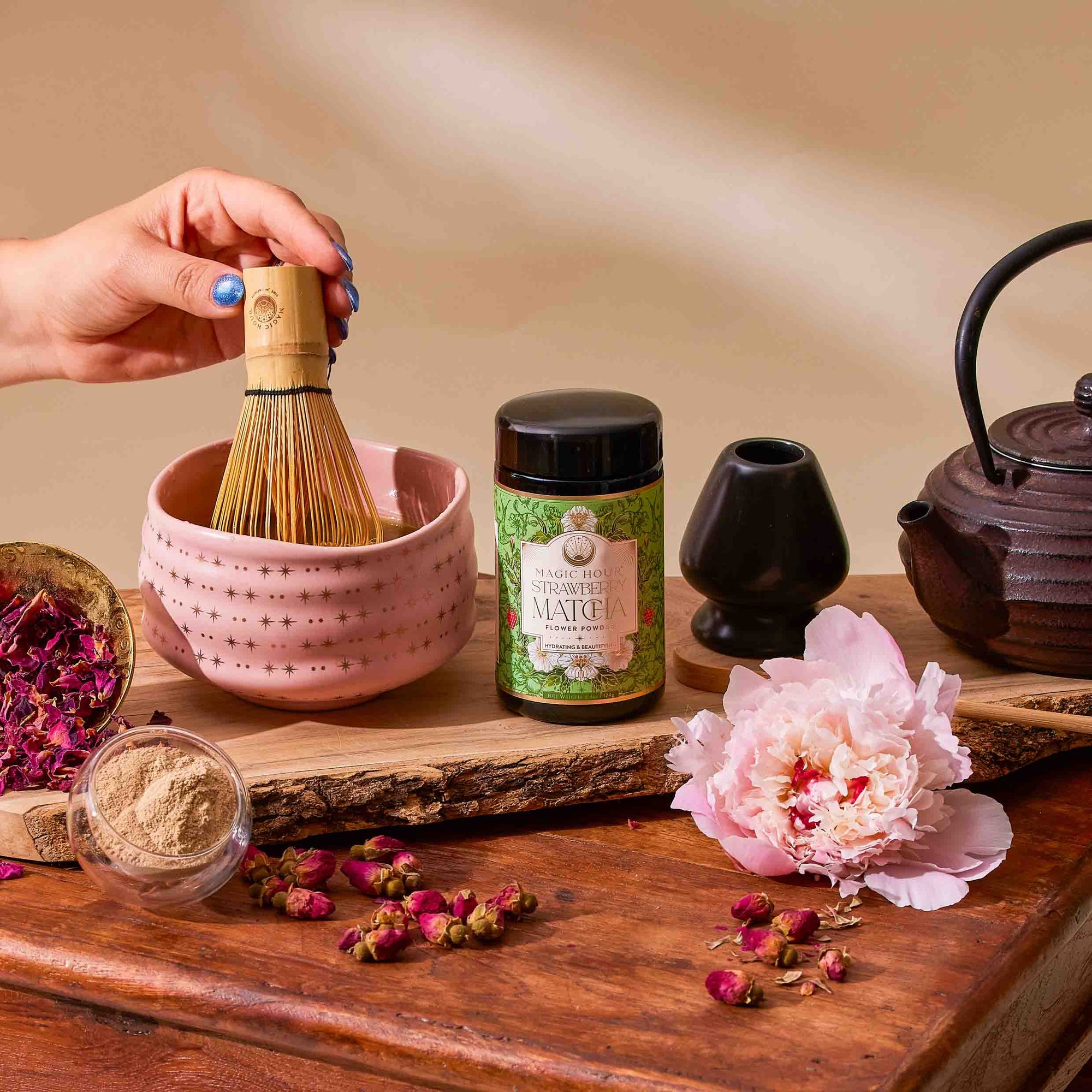 A hand whisks ceremonial matcha from the Flower Powder: Matcha Bundle in a pink bowl, with the bundle’s powder jar, teapot, scoop, dried rose petals, peony flower, and wooden tray arranged nearby.