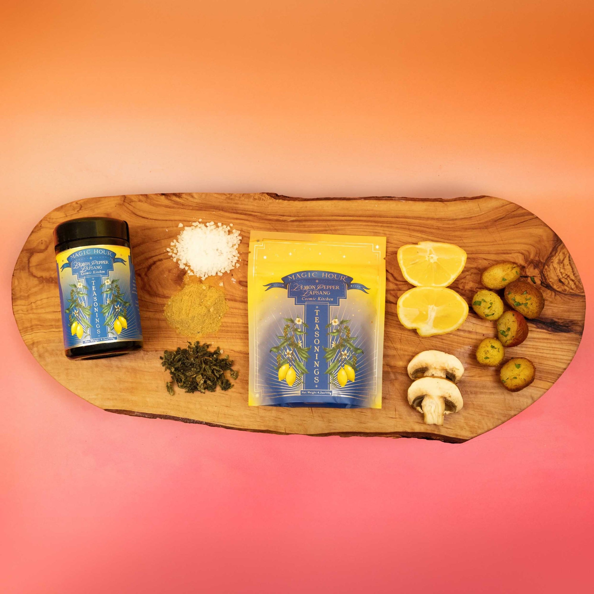 A wooden board displays a Teasoning: Lemon Pepper Lapsang pouch, seasoning jar, coarse salt, green herbs, lemon halves, potato slices, mushroom slices, and cucumber slices against a pink-orange gradient background.