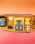 A wooden board displays a Teasoning: Lemon Pepper Lapsang pouch, seasoning jar, coarse salt, green herbs, lemon halves, potato slices, mushroom slices, and cucumber slices against a pink-orange gradient background.