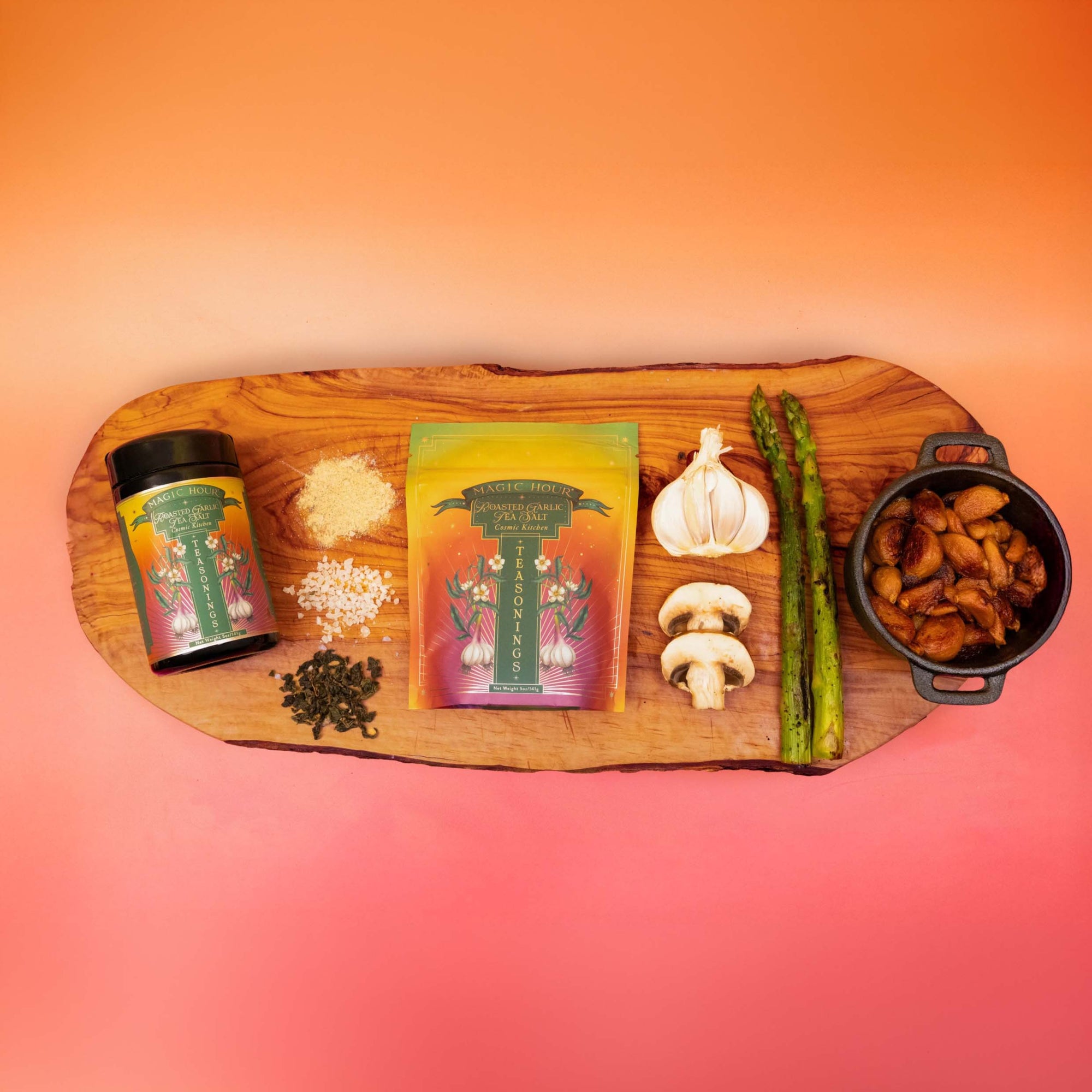 A wooden board displays Teasoning: Roasted Garlic Tea Salt, a spice jar, grated cheese, rice, peppercorns, mushrooms, asparagus, garlic, and a small pot of cooked potatoes—all set against an orange gradient background.