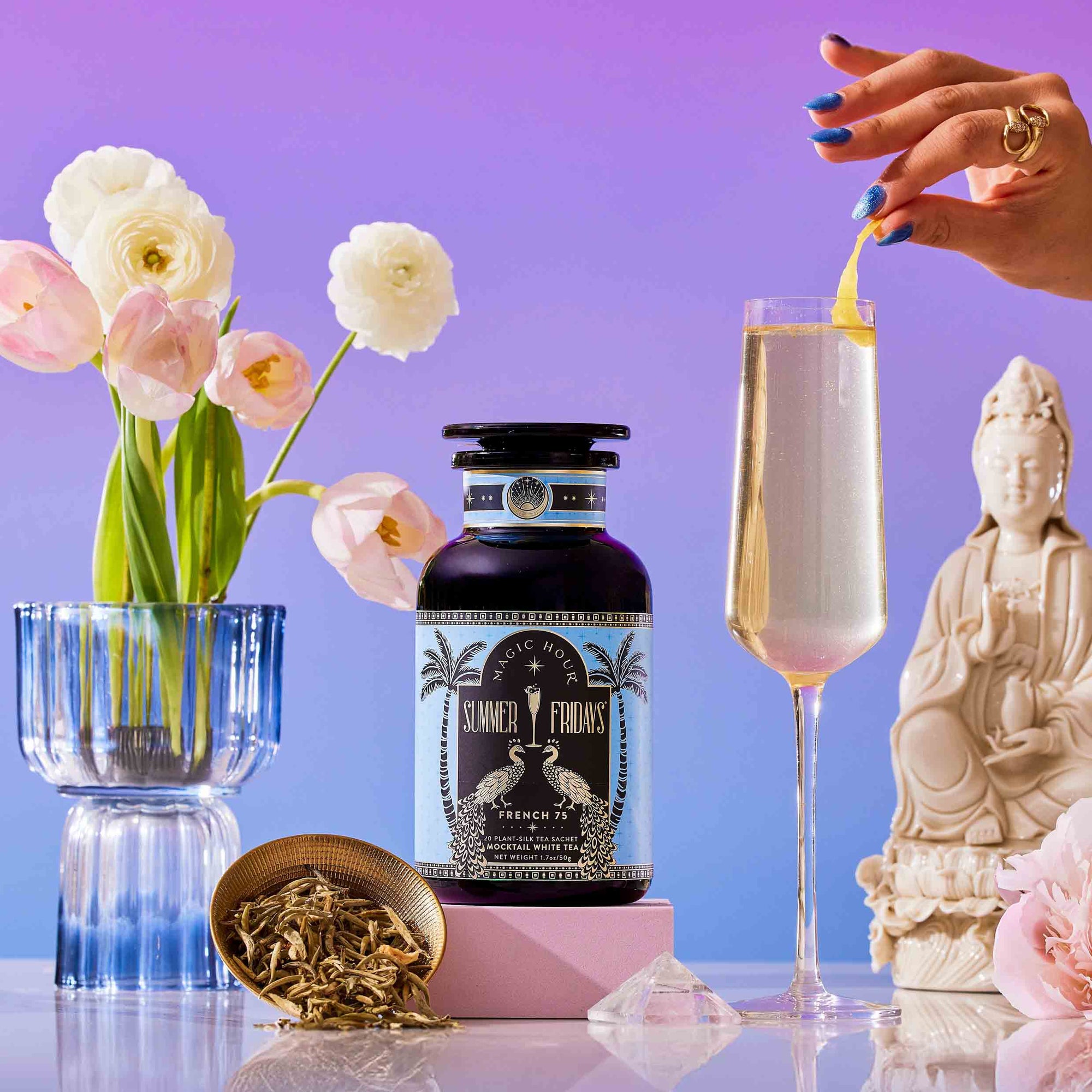 A hand garnishes a champagne flute with lemon twist beside a Summer Fridays: Wellness Mocktail Tea Bundle. Pink flowers in a blue vase, loose tea, and a small statue sit on a pastel surface against a purple gradient background.