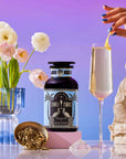 A hand garnishes a champagne flute with lemon twist beside a Summer Fridays: Wellness Mocktail Tea Bundle. Pink flowers in a blue vase, loose tea, and a small statue sit on a pastel surface against a purple gradient background.