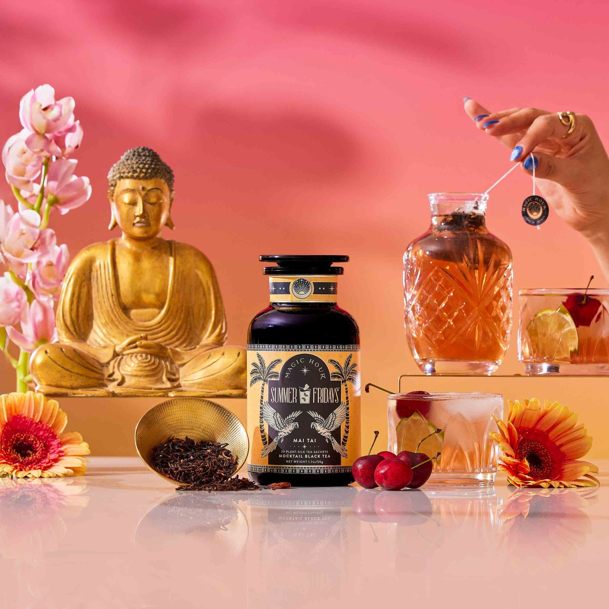 A bottle labeled “Summer Fridays™: Mai Tai” sits on a glossy surface with tea leaves, cherries, and flowers nearby. In the background, a golden Buddha statue and a hand stir a zero-proof cocktail under a pink-orange sky.