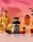 A bottle from our Summer Fridays: Wellness Mocktail Tea Bundle rests on a reflective surface beside a gold Buddha statue, iced tea, flowers, cherries, loose tea leaves, and a hand stirring a drink against a vibrant pink-orange backdrop.