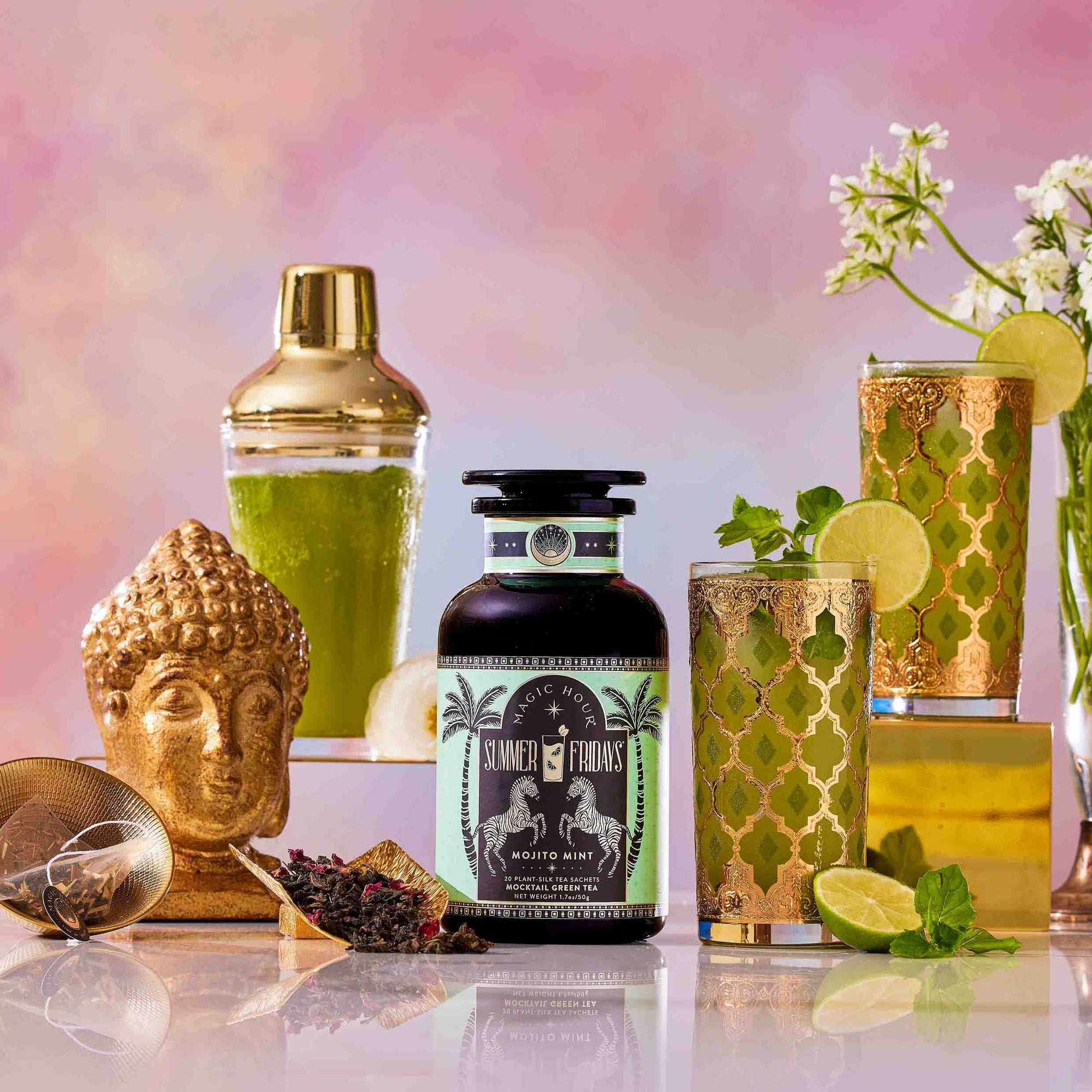 A bottle of Summer Fridays™: Mojito Mint stands among a cocktail shaker, ornate zero-proof cocktails, lime slices, a golden Buddha head, a tea infuser, and flowers on a vibrant background.