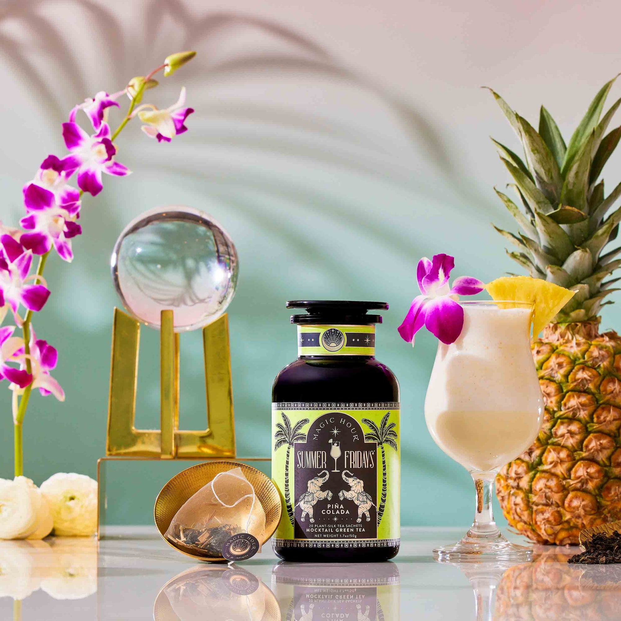 The Summer Fridays: Wellness Mocktail Tea Bundle sits next to a piña colada mocktail in a glass garnished with pineapple and an orchid, surrounded by a pineapple, botanical tea infuser, flowers, and a crystal ball on a stand.