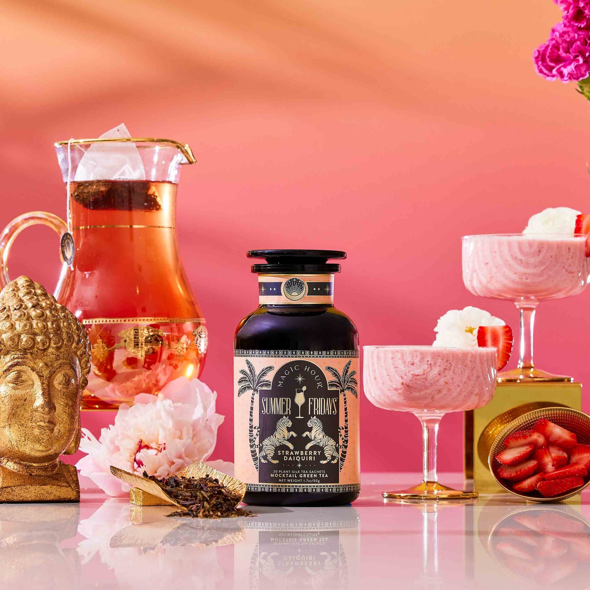 A bottle of Summer Fridays™: Strawberry Daiquiri, a summer tea blend, stands on a reflective surface beside a glass pitcher of tea, two glasses of strawberry mousse, a gold Buddha head, pink flowers, and sliced strawberries against a pink gradient background.