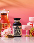 A bottle of Summer Fridays™: Strawberry Daiquiri, a summer tea blend, stands on a reflective surface beside a glass pitcher of tea, two glasses of strawberry mousse, a gold Buddha head, pink flowers, and sliced strawberries against a pink gradient background.