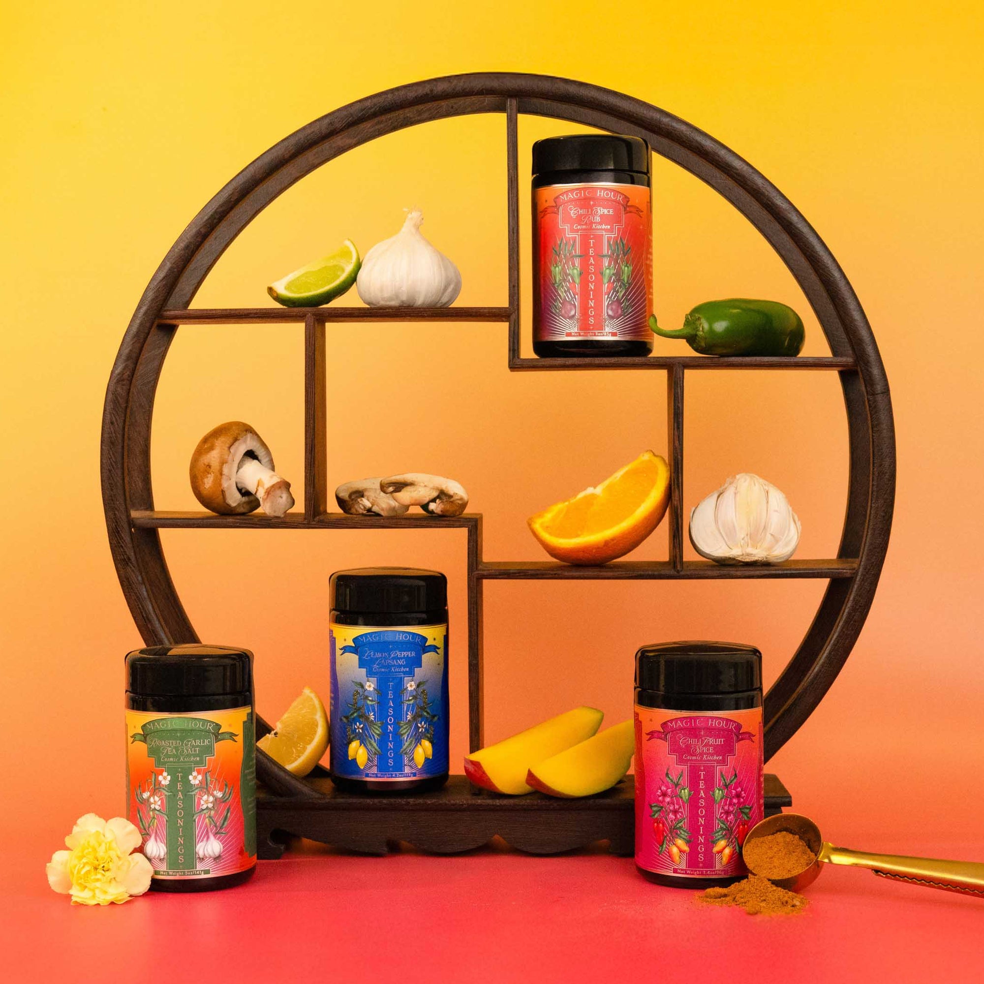 The Teasoning Bundle—tea-infused botanical seasonings—is showcased in four colorful jars on a round wooden shelf with lime, garlic, jalapeño, mushrooms, and orange slices. A gold spoon and flowers complete the vibrant display.