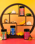 The Teasoning Bundle—tea-infused botanical seasonings—is showcased in four colorful jars on a round wooden shelf with lime, garlic, jalapeño, mushrooms, and orange slices. A gold spoon and flowers complete the vibrant display.