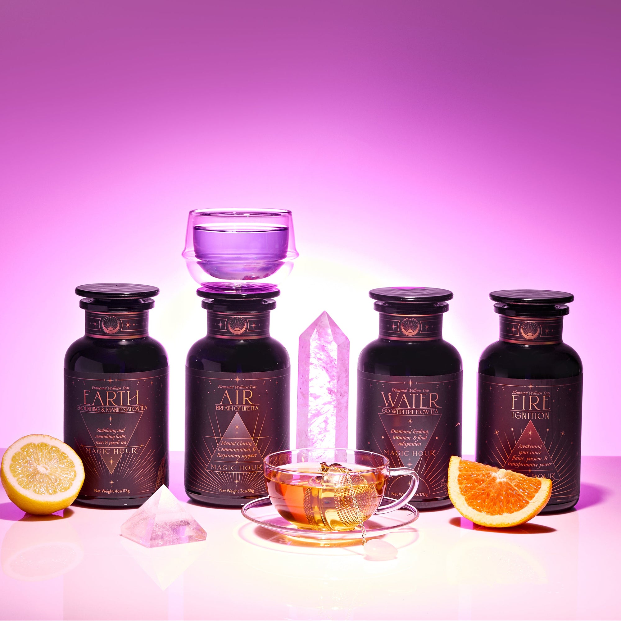The Elemental Alchemy Master Collection features four dark glass bottles labeled Earth, Air, Water, Fire, a glowing pink crystal, a glass teacup with purple Elemental Alchemy tea, lemon and orange slices, set against a pink gradient background.