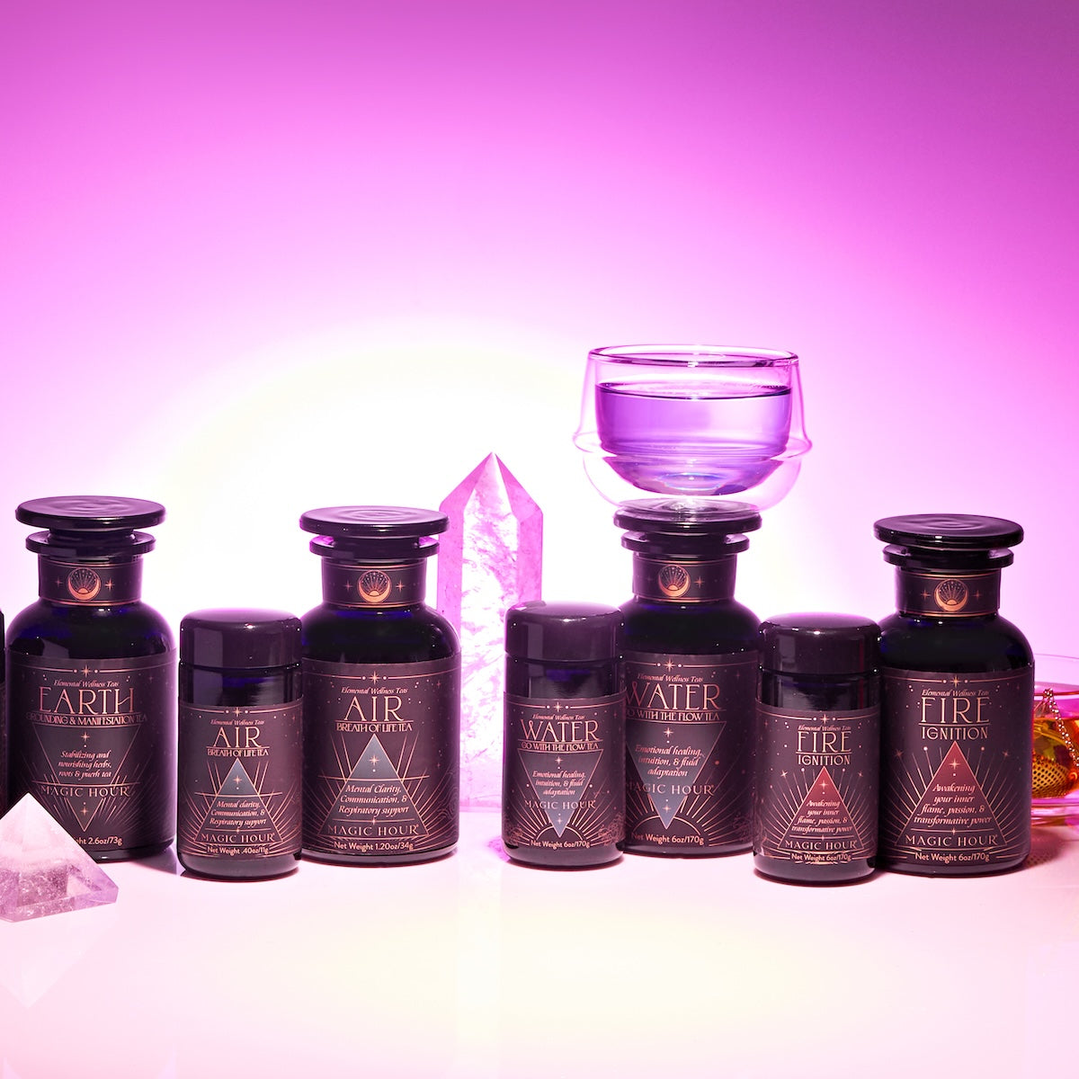 A Monthly Tea Subscription Box with six dark bottles featuring geometric labels—Earth, Air, Water, or Fire—rests on a white surface near purple crystals and a glass cup against a pink and purple gradient background.
