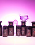 A Monthly Tea Subscription Box with six dark bottles featuring geometric labels—Earth, Air, Water, or Fire—rests on a white surface near purple crystals and a glass cup against a pink and purple gradient background.