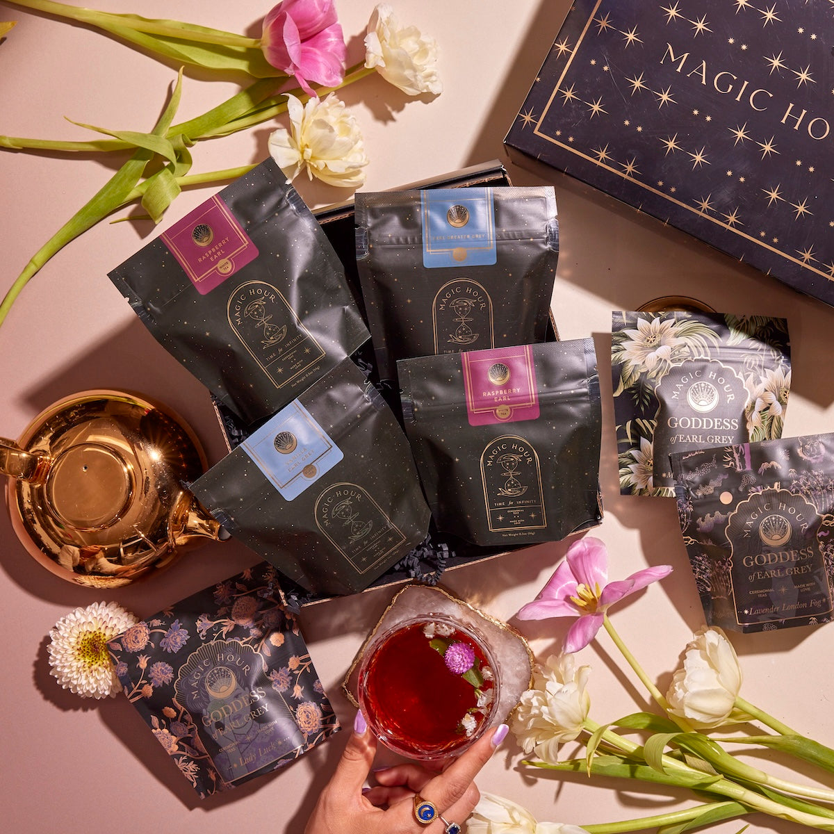 Flat lay showcasing a tea set with several bags of "Magic Hour" branded tea, including the bergamot-infused "Goddess" blend, beautifully arranged alongside fresh flowers. A pot of gold-colored tea and teabags from the Earl Grey Sampler Set add to the display. Additionally, a hand holds a glass of red tea garnished with flowers and a "Magic Hour" box.