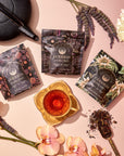 Top-down view of a luxurious tea setup featuring The Goddess of Earl Set from Magic Hour, including three tea pouches, a black teapot, and a golden cup filled with tea. The scene is adorned with orchid flowers, lavender sprigs, a Gold Luxe Loose Leaf Tea Strainer with loose tea leaves, and a white chrysanthemum flower.