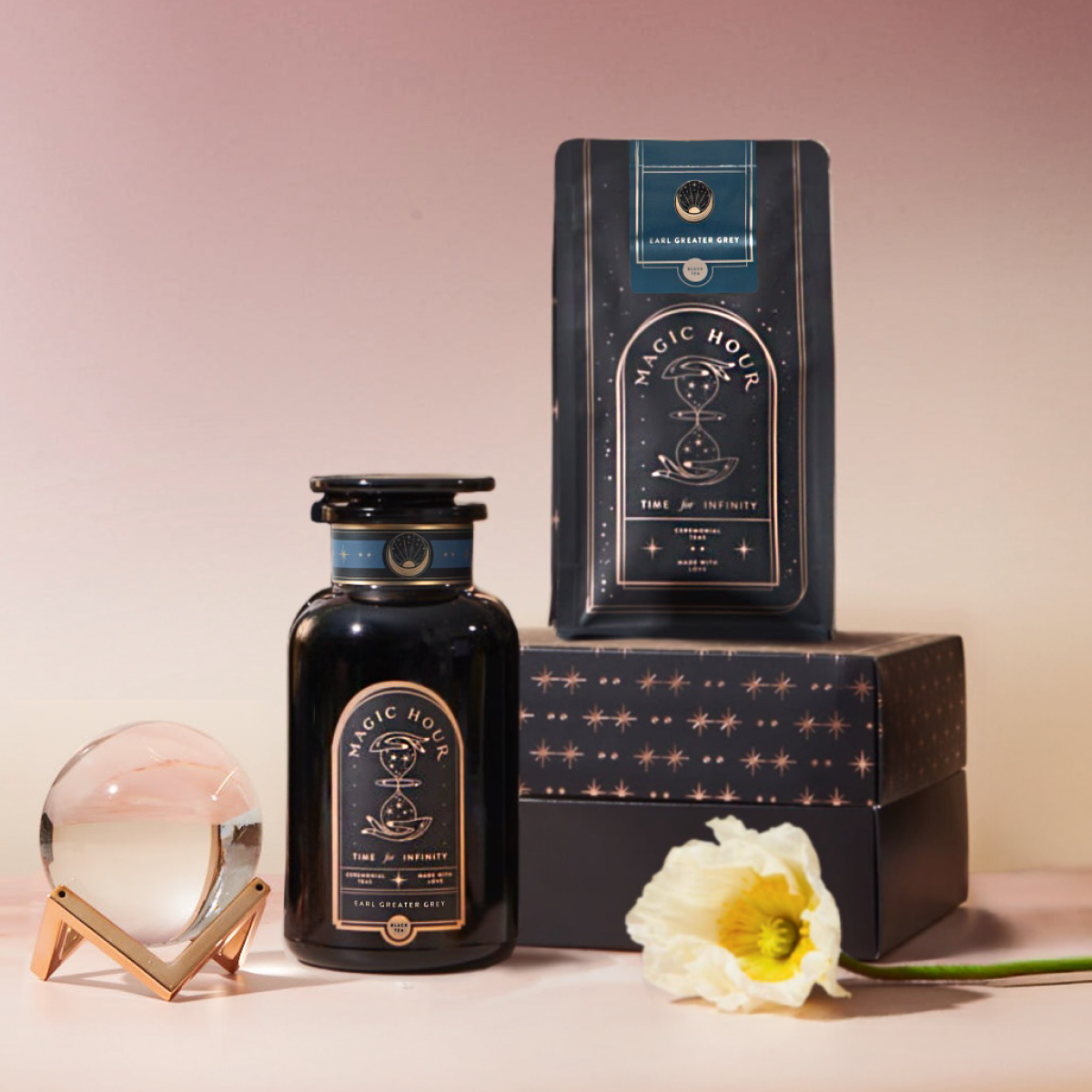 A black jar and matching tea bag packet labeled "Earl Greater Grey Starter Kit" sit on a star-patterned box with a white flower and glass sphere on a rose-gold stand, set against a pink gradient background.