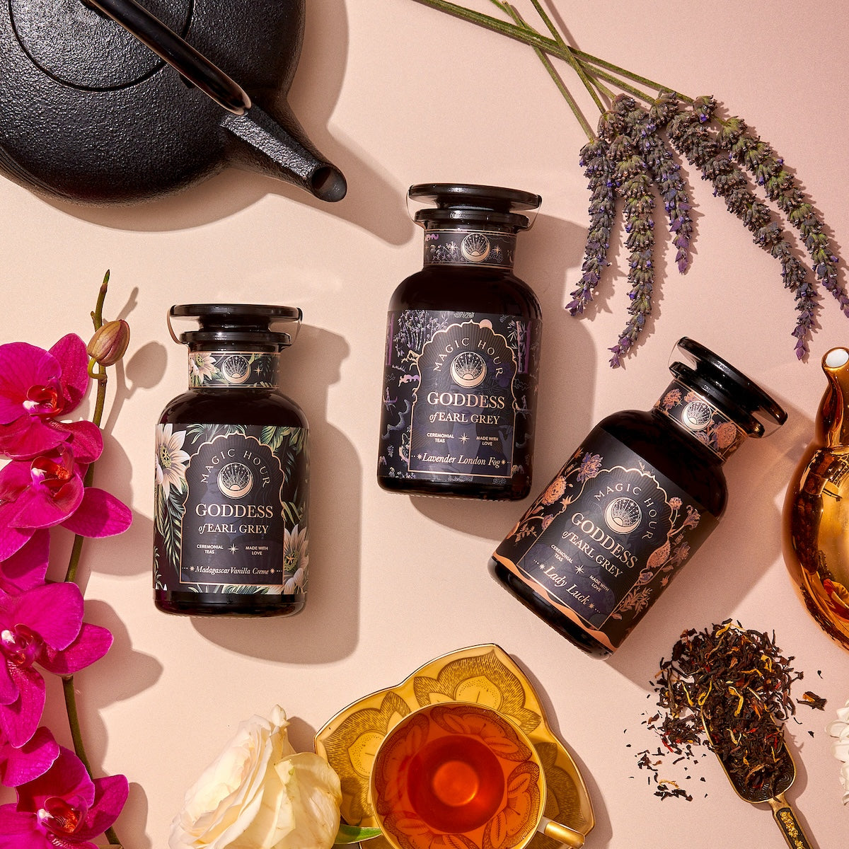 A flat lay photo showcases three glass bottles of The Goddess of Earl Set by Magic Hour, a goddess-themed Earl Grey tea collection. The bottles are beautifully displayed alongside a teapot, a filled teacup, loose tea leaves, the Gold Luxe Loose Leaf Tea Strainer, fresh lavender, and vibrant pink orchids on a beige surface.